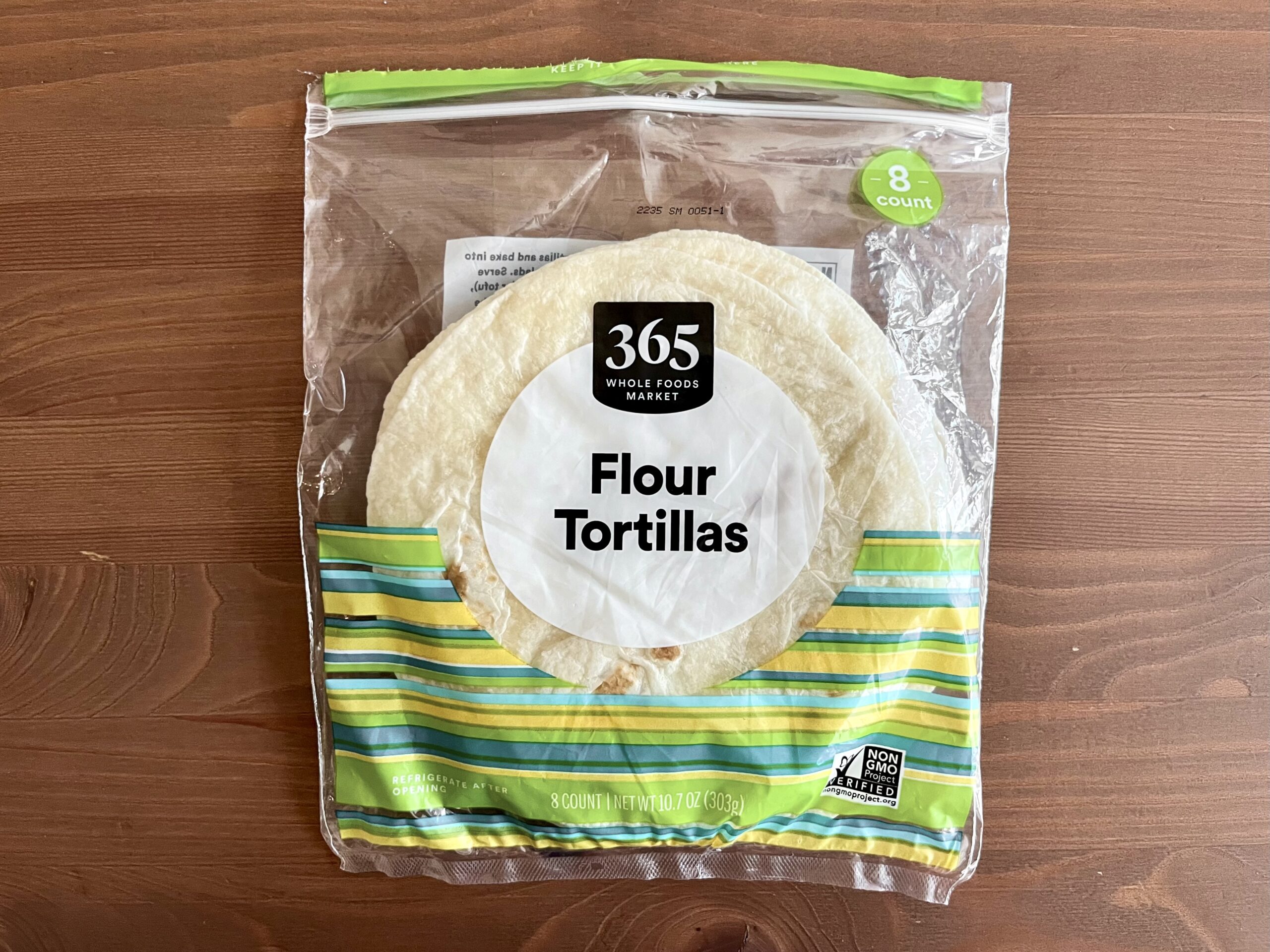 A plastic package of 365 Whole Foods Market Flour Tortillas sits on a wooden surface. The package is green and yellow striped at the bottom and contains 8 tortillas.