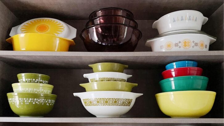 A shelf displays colorful vintage bowls and dishes, including yellow, green, white, red, blue, and purple Pyrex bowls, some with floral or geometric patterns, neatly stacked in rows.