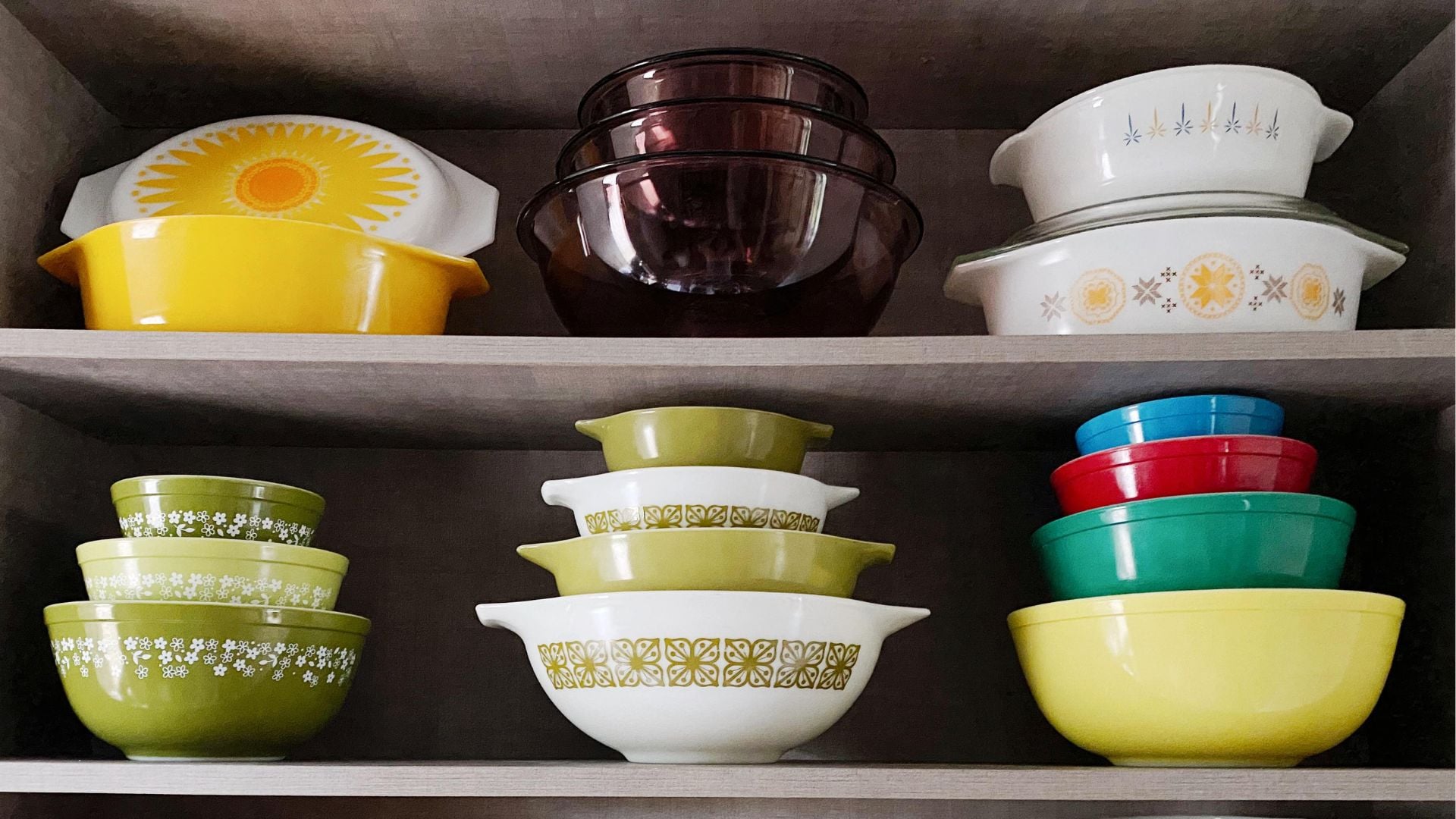 A shelf displays colorful vintage bowls and dishes, including yellow, green, white, red, blue, and purple Pyrex bowls, some with floral or geometric patterns, neatly stacked in rows.