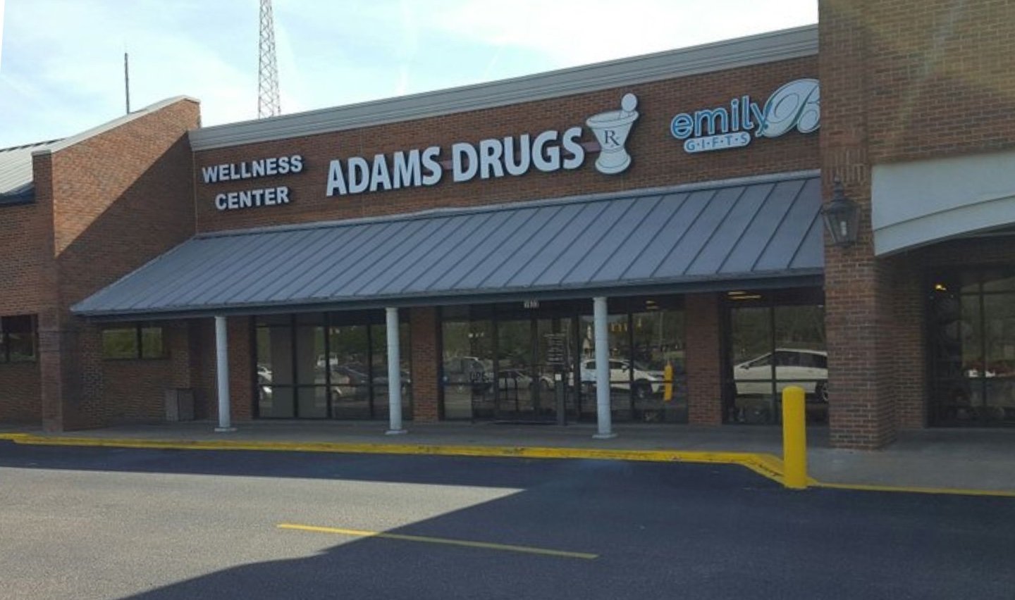 A brick storefront with signs reading “WELLNESS CENTER,” “ADAMS DRUGS” with a mortar and pestle icon, and “emily GIFTs.” Large windows and a metal awning are above the sidewalk and parking lot.