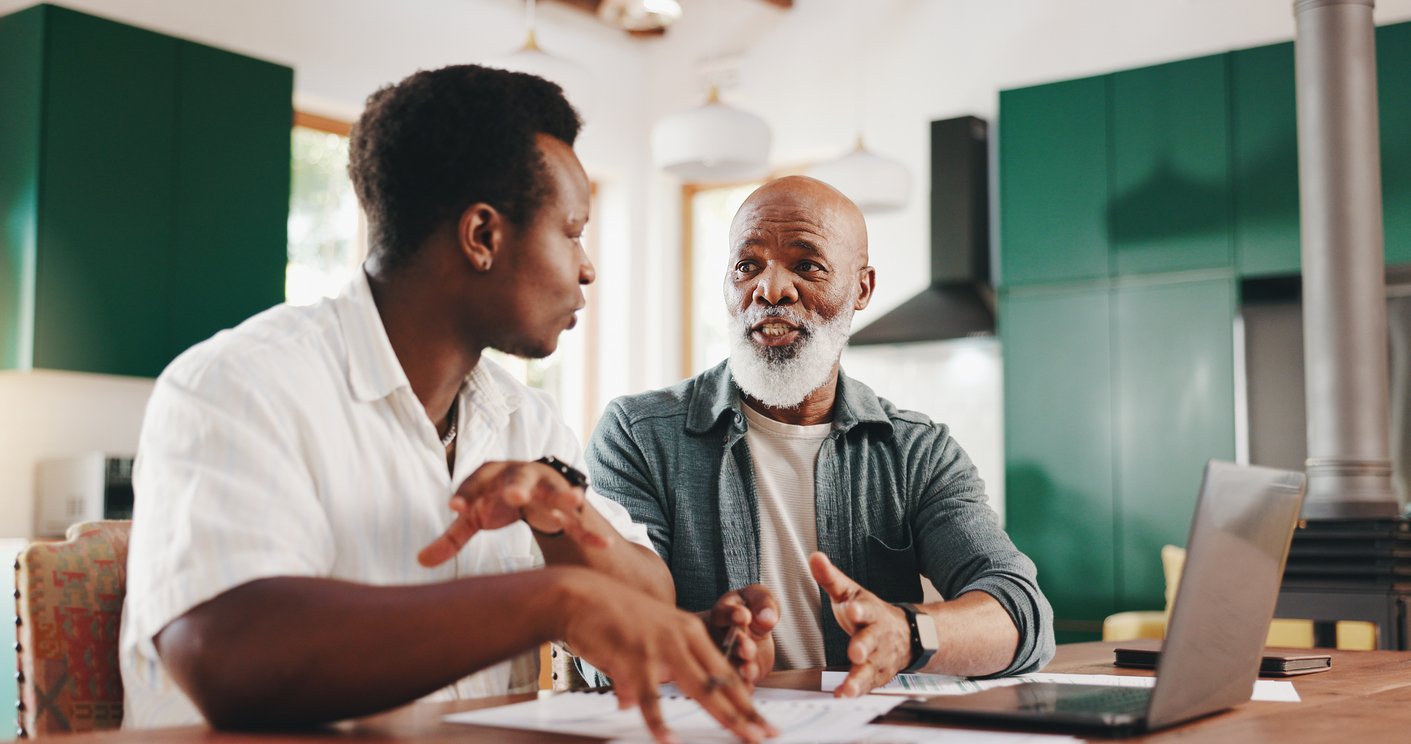 Two men sit at a table in a kitchen, engaged in conversation. Papers and a laptop are on the table in front of them. The older man gestures as he speaks, while the younger man listens attentively.