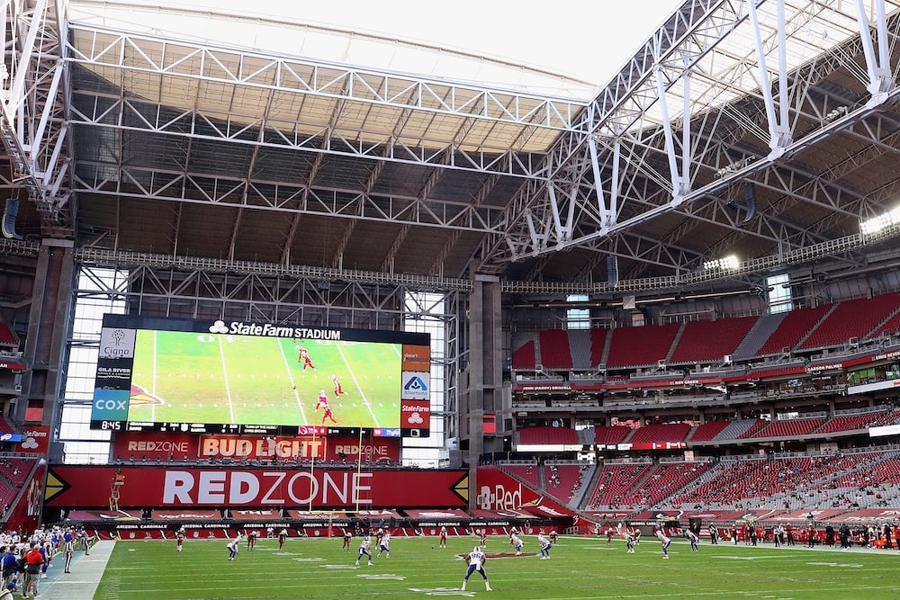 A football game takes place inside State Farm Stadium, with players on the field and a large video screen showing the game action. The stands have sparse attendance, and the stadium roof is partially open.