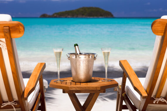 Two wooden lounge chairs face a turquoise ocean with an island in the distance. A small table between them holds a champagne bucket with a bottle and two filled champagne flutes. The scene is bright and tropical.