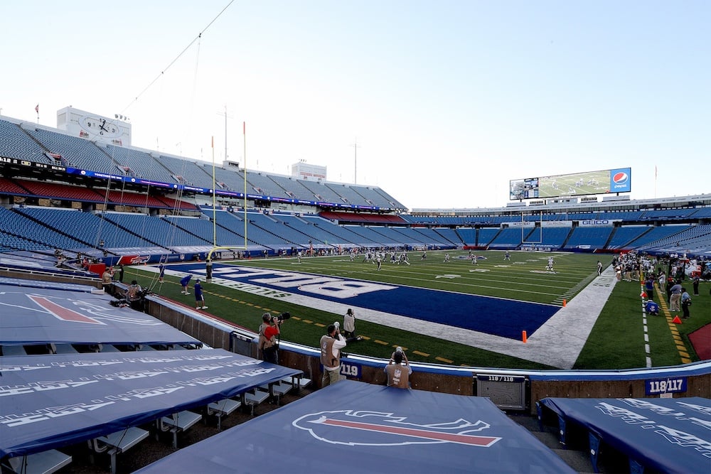 A view of an empty football stadium with players and staff on the field. The end zone features the Buffalo Bills logo, and the stands are mostly empty with some people near the sideline.