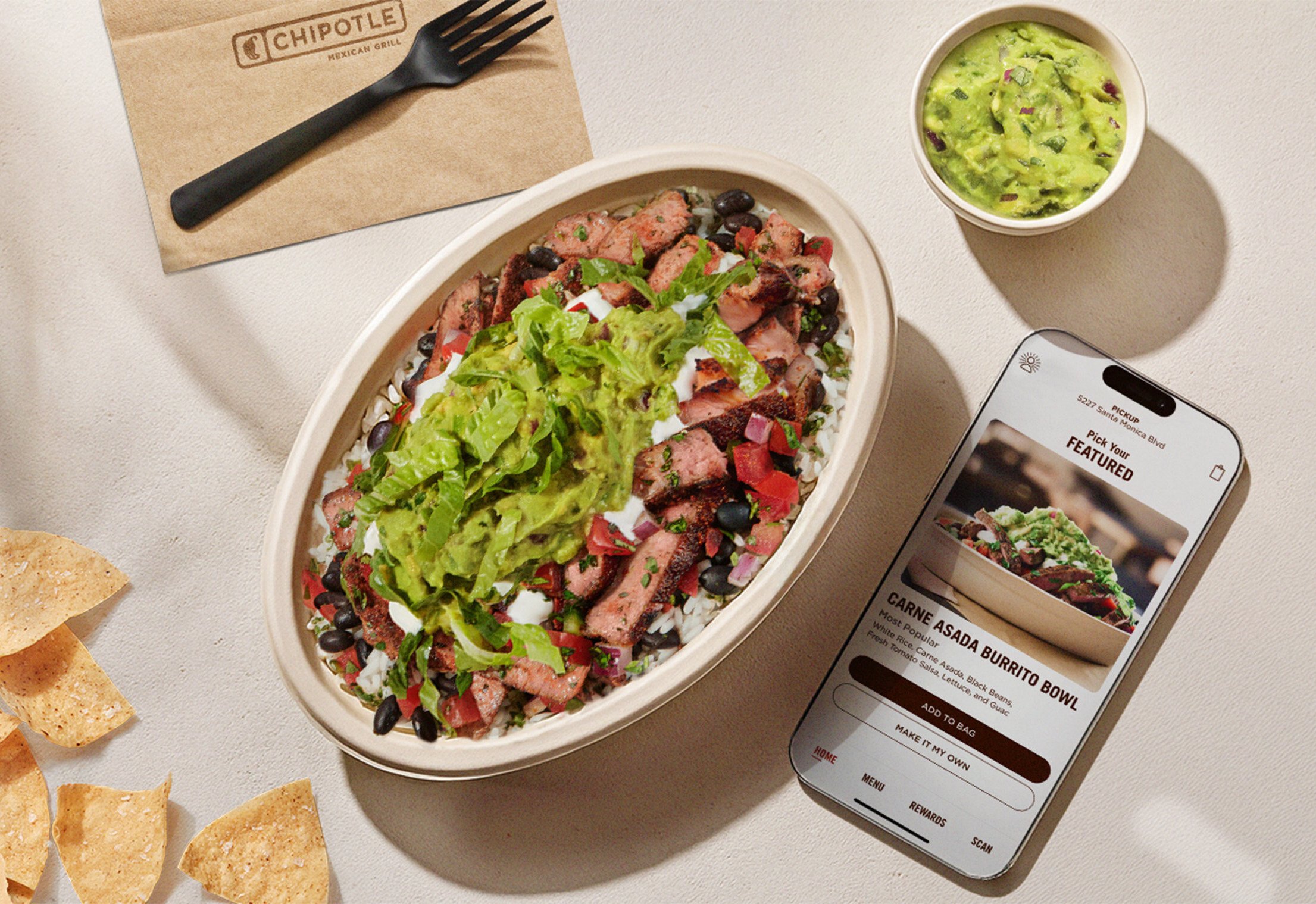 A Chipotle burrito bowl with steak, rice, beans, salsa, and guacamole sits next to tortilla chips, a cup of guacamole, a Chipotle napkin, and a phone displaying the Carne Asada Burrito Bowl on the Chipotle app.
