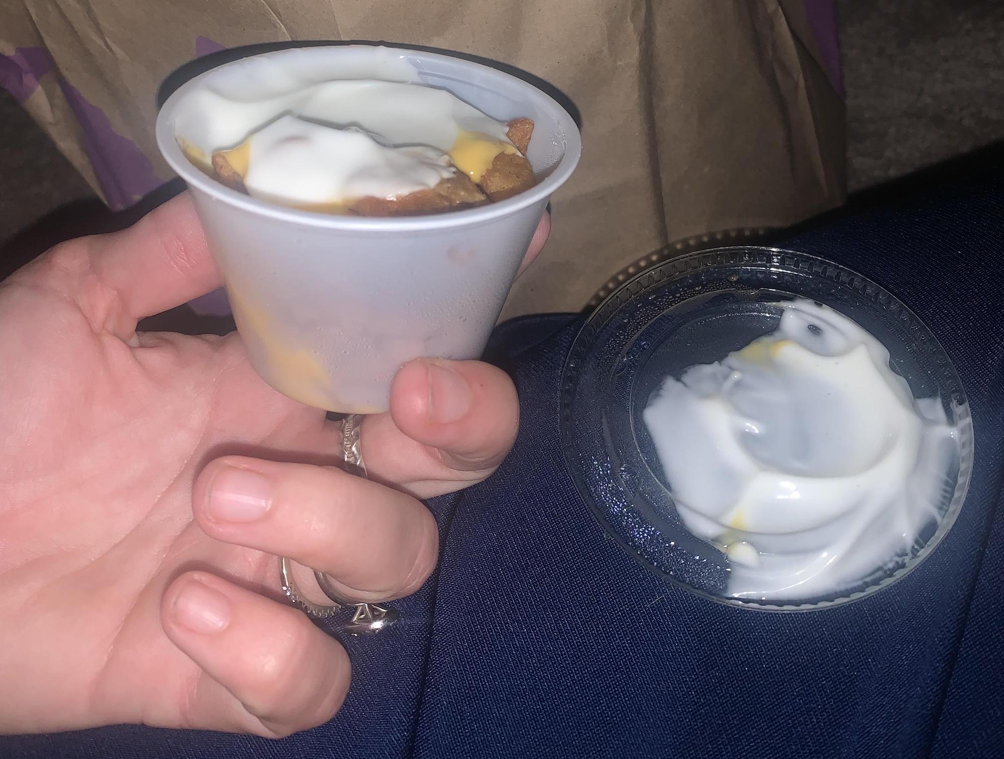 A hand holds a small plastic cup filled with nachos topped with cheese and sour cream. The clear lid, with remnants of sauce, rests nearby on a blue fabric surface. Taco Bell Cheesy Fiesta Potatoes