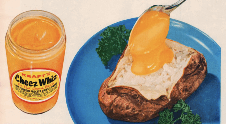 A spoon pours bright orange Cheez Whiz sauce—an infamous example of American food—onto a baked potato on a blue plate, garnished with parsley. An open jar of Cheez Whiz sits next to the plate.