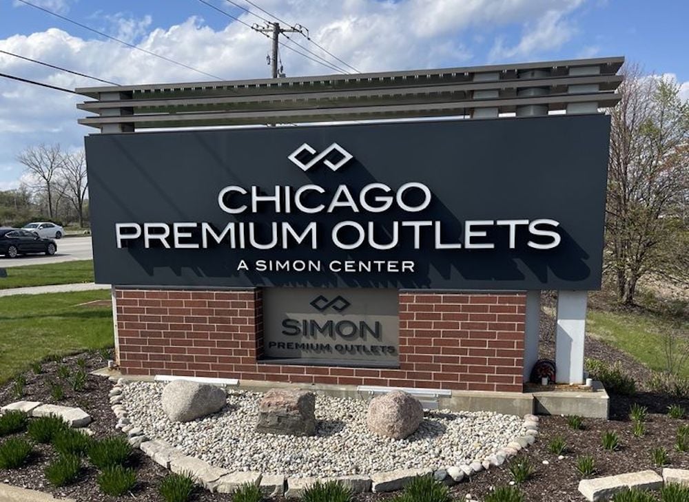 A large sign for Chicago Premium Outlets, a Simon Center, stands on a brick and stone base with landscaping, against a background of trees and a partly cloudy sky.