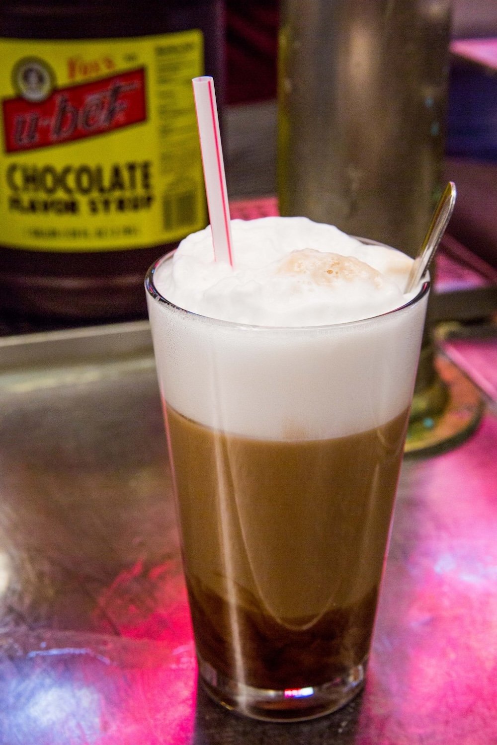 A glass filled with an egg cream, featuring a thick layer of white foam on top and a red-striped straw, sits on a counter. A spoon is also in the glass, and a bottle of chocolate syrup is in the background.