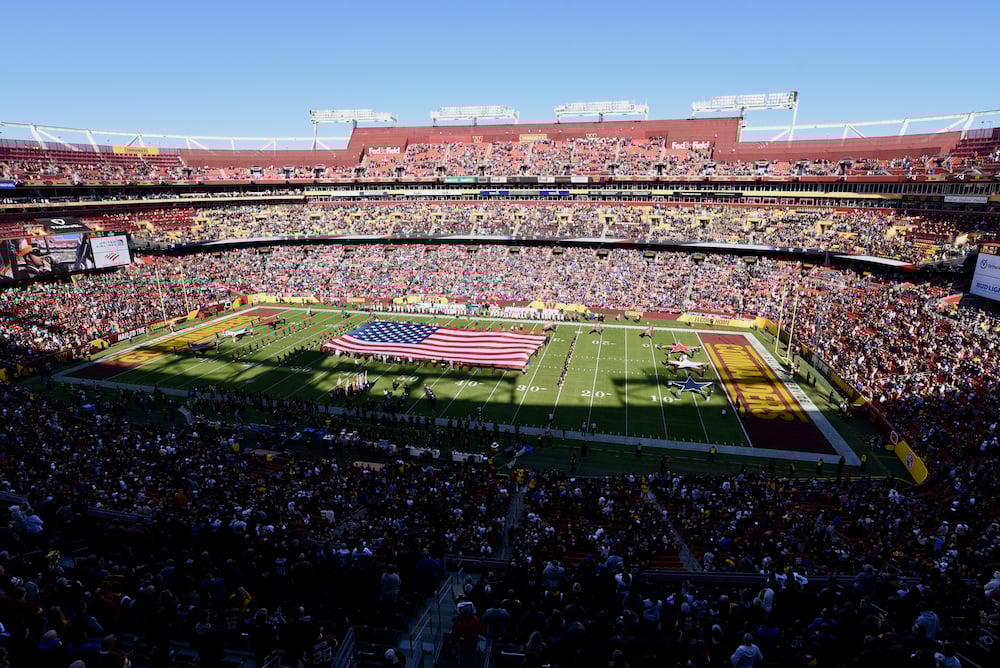 A large American flag is displayed across the football field during a pre-game ceremony at a packed stadium under a clear blue sky. Spectators fill the stands, and players and personnel stand on the sidelines.