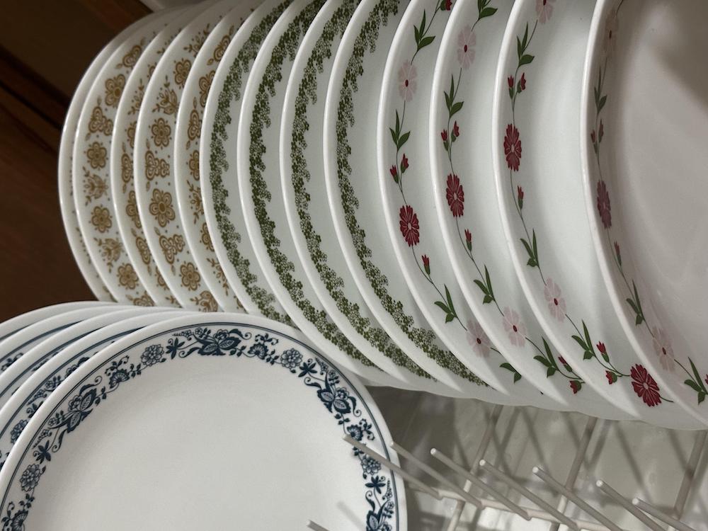 Several white plates with different floral and patterned borders—blue, gold, green, and red—are arranged vertically in a dish rack for drying.