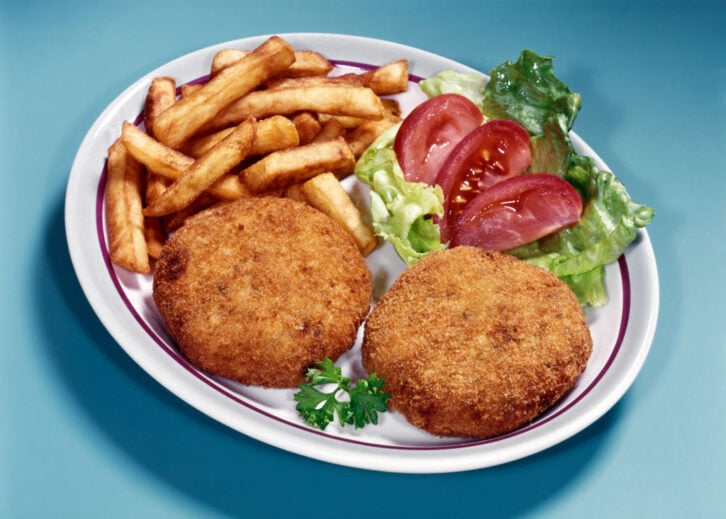 A plate with two breaded crab cakes, a serving of French fries, lettuce, tomato slices, and a sprig of parsley on a light blue background.