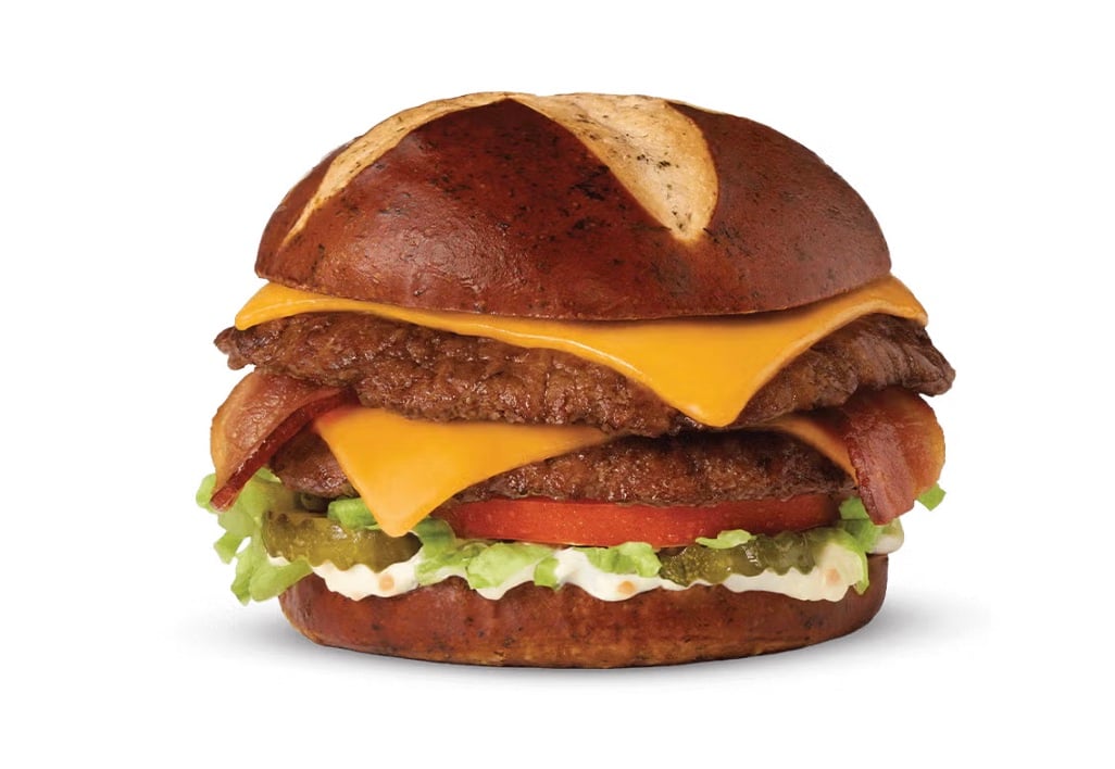 A new fast-food favorite: a hamburger with a pretzel bun, two beef patties, cheddar cheese, bacon, lettuce, tomato, pickles, and mayonnaise, all served against a white background.