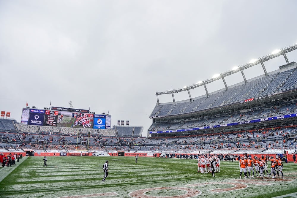 A football stadium with a partially filled crowd; players from two teams gather on the snowy field near the sideline. A large scoreboard and overcast sky are visible in the background.