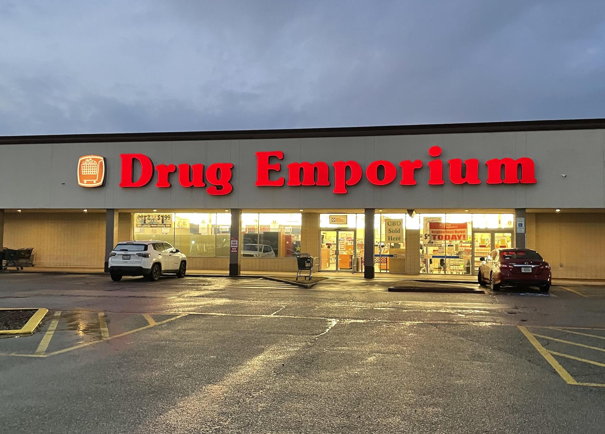 A brightly lit Drug Emporium store at dusk with two parked cars in front, glowing red signage, and wet pavement reflecting the store lights.