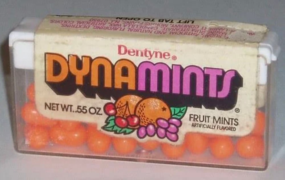 A clear plastic box of Dentyne DynaMints fruit mints featuring an illustrated label with oranges and grapes, showing small round orange mints inside.