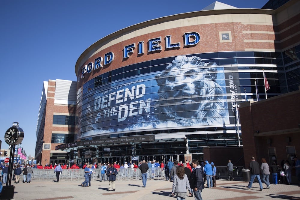 People gather outside Ford Field, a large stadium with a mural of a roaring lion and the words "DEFEND THE DEN" across the windows. A clock and several flags are visible in the plaza.