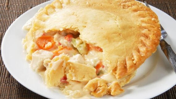 A creamy chicken pot pie with a golden, flaky crust sits on a white plate. A portion is missing, revealing chunks of chicken, carrots, and vegetables in a thick sauce.