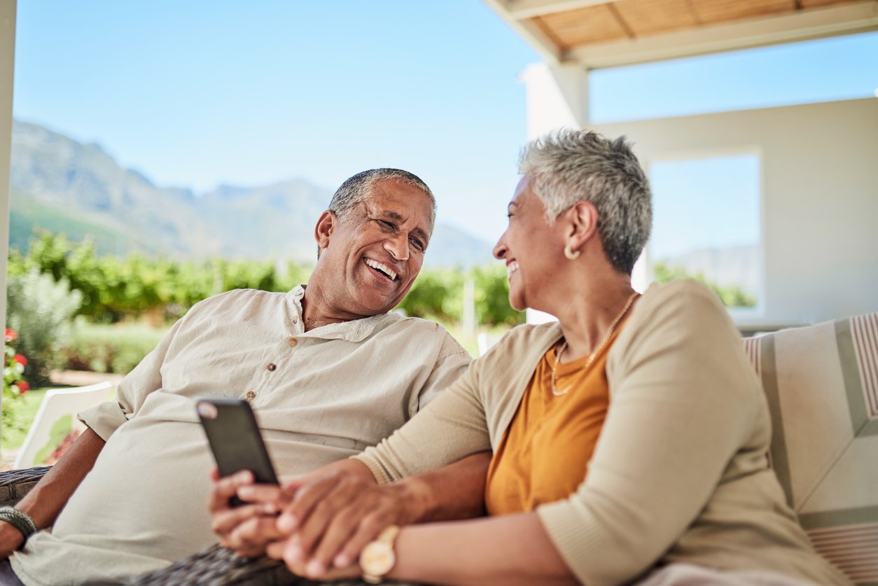 A smiling older couple sit together outdoors on a shaded patio, holding hands and looking at each other. One holds a smartphone, and mountains with lush greenery are visible in the background.