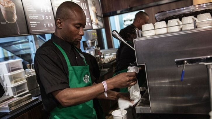 A barista wearing a green apron works behind the counter at a coffee shop, preparing a drink next to a large espresso machine. Shelves with cups and a menu board are visible in the background.