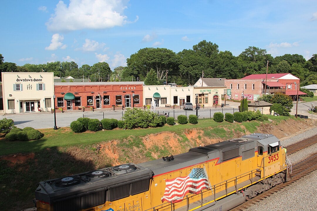 A yellow Union Pacific train engine passes in front of a small-town street with historic brick buildings, shops, and greenery under a partly cloudy blue sky.
