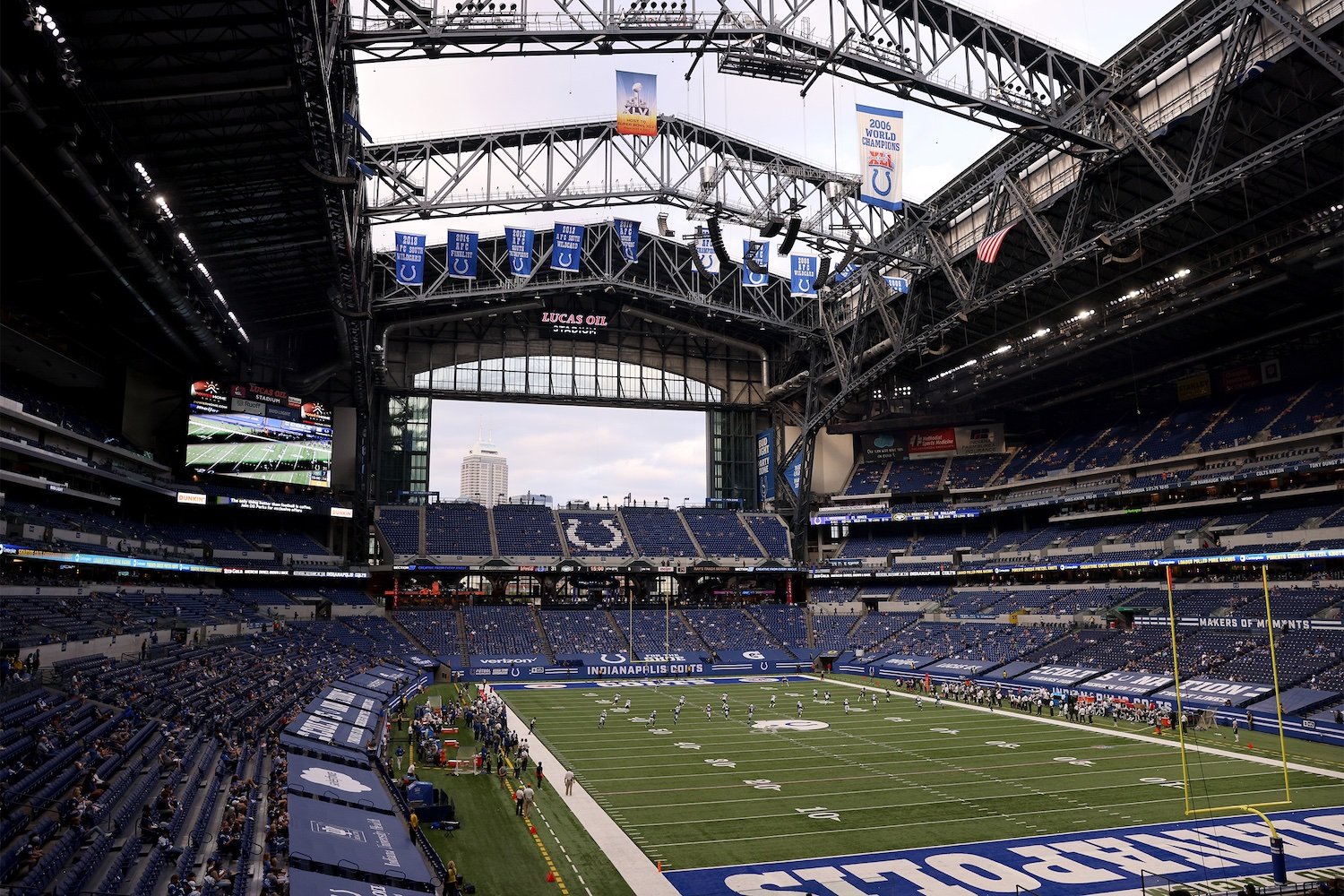 A wide view of an indoor football stadium with a partially open roof, showing the field, seats, scoreboard, and banners; some fans are scattered throughout the stands.
