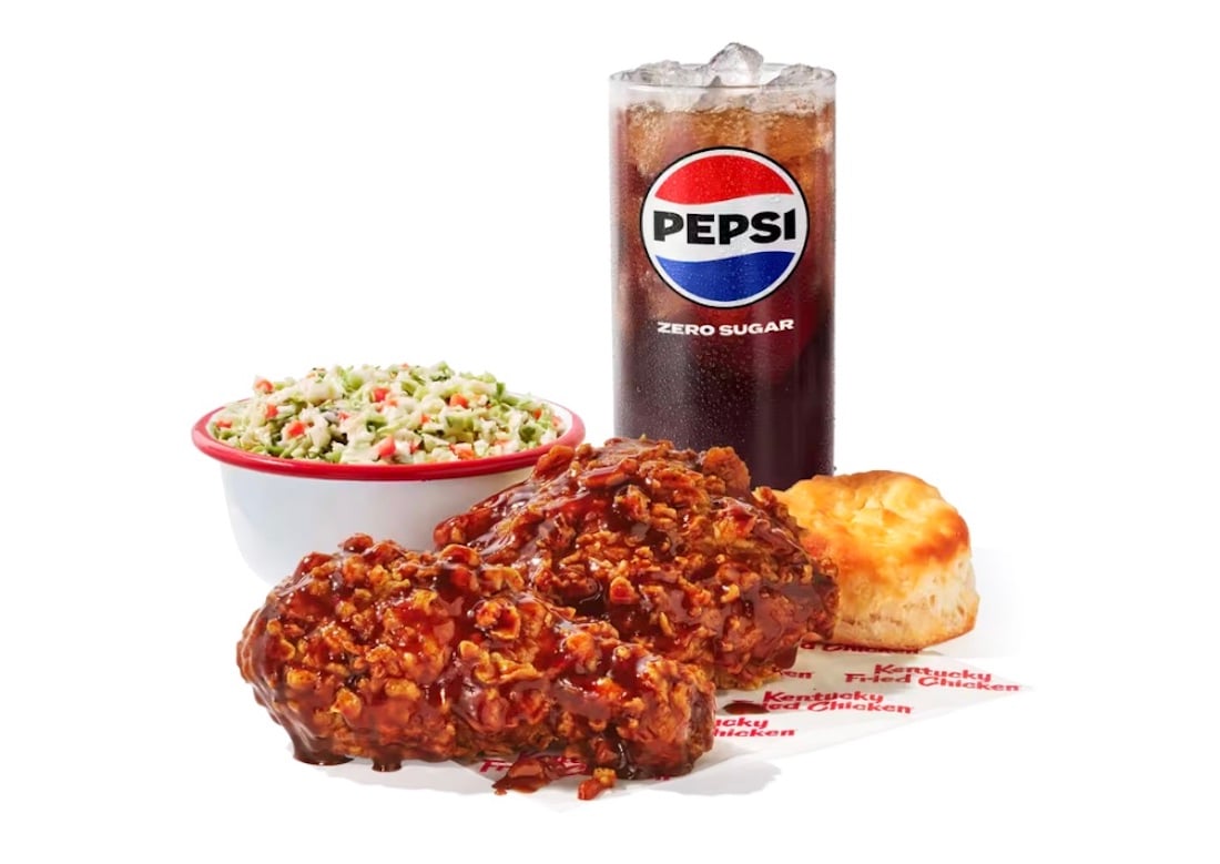 Enjoy this new fast-food meal: two pieces of saucy fried chicken, a biscuit, creamy coleslaw, and a glass of Pepsi Zero Sugar with ice, all arranged on a white background.