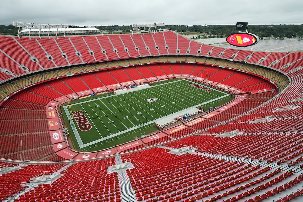 An empty football stadium with red seats and a green field marked with "CHIEFS" in the end zones, viewed from above under a cloudy sky. The large stadium is surrounded by a scoreboard and trees in the distance.