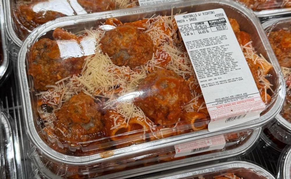 Plastic containers filled with pasta and meatballs in tomato sauce, topped with shredded cheese, are stacked on a shelf with a white label showing product details and price.