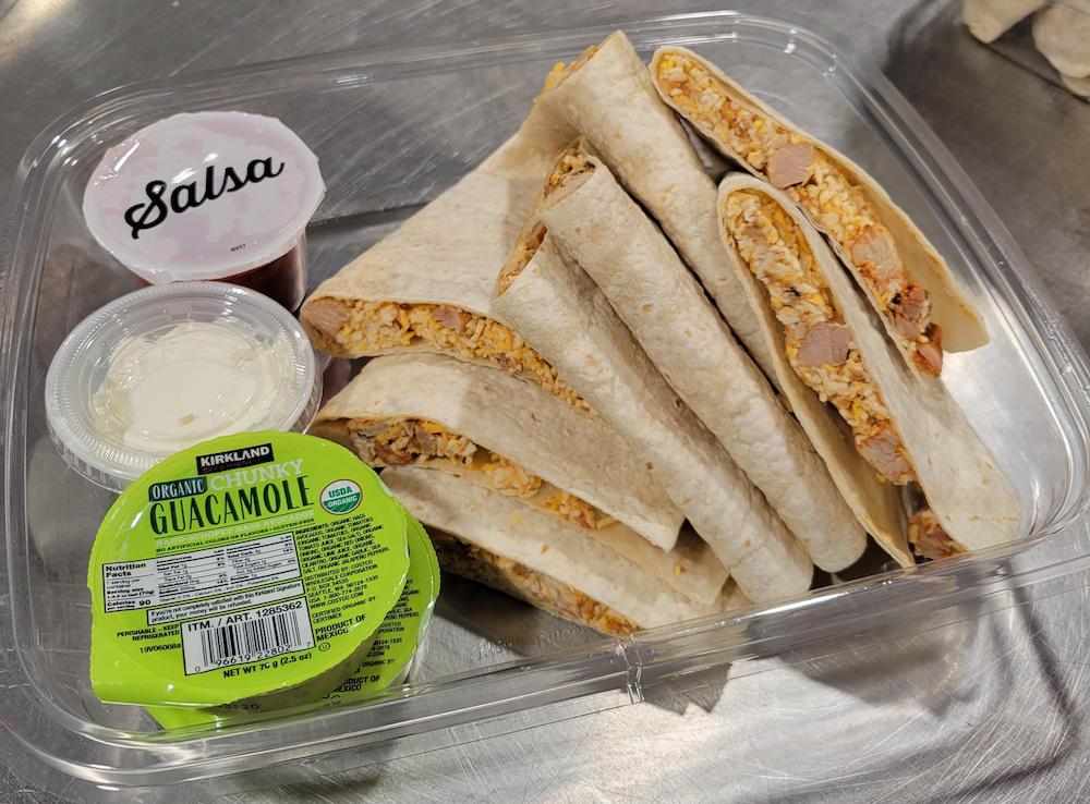 A plastic container with sliced wraps filled with rice and sausage, served with individual cups of salsa, sour cream, and organic chunky guacamole on a stainless steel surface. Kirkland Signature Chicken Quesadillas.