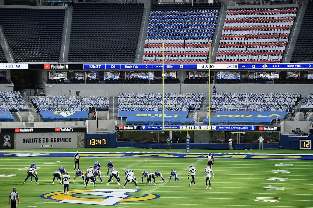 Football teams line up on a field in a mostly empty stadium. The seats behind them are covered to display an American flag pattern, while banners read "BUILT FOR LA." A scoreboard and digital play clock are also visible.