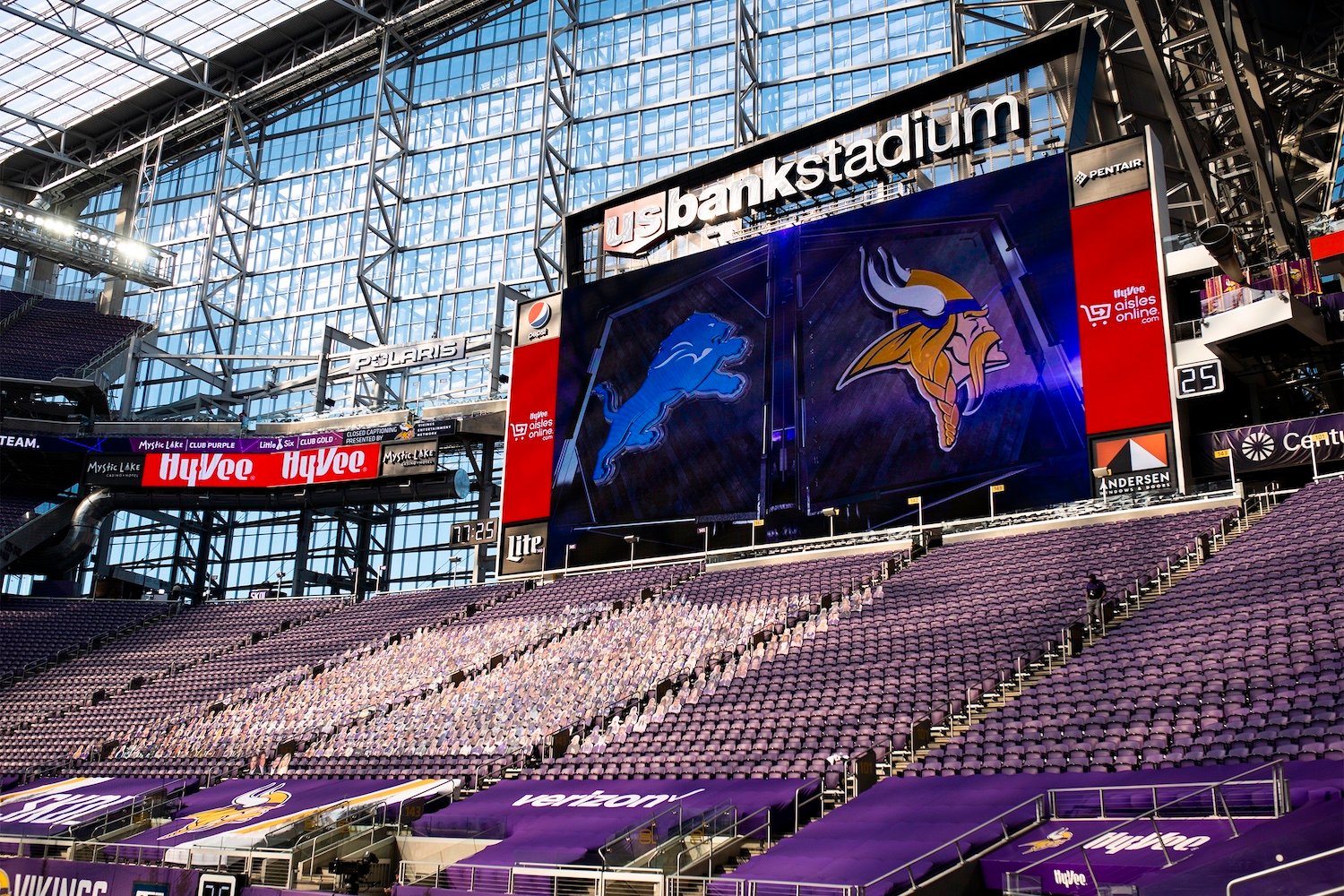 A football stadium with purple seats, large digital screens showing Detroit Lions and Minnesota Vikings logos, and a US Bank Stadium sign above. The stands are mostly empty.