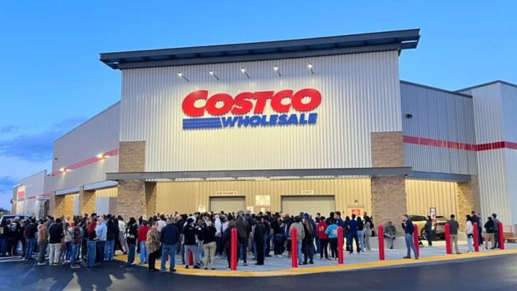 A large crowd of people waits in line outside a Costco Wholesale store entrance at dusk, beneath a prominent Costco sign on the building’s facade.