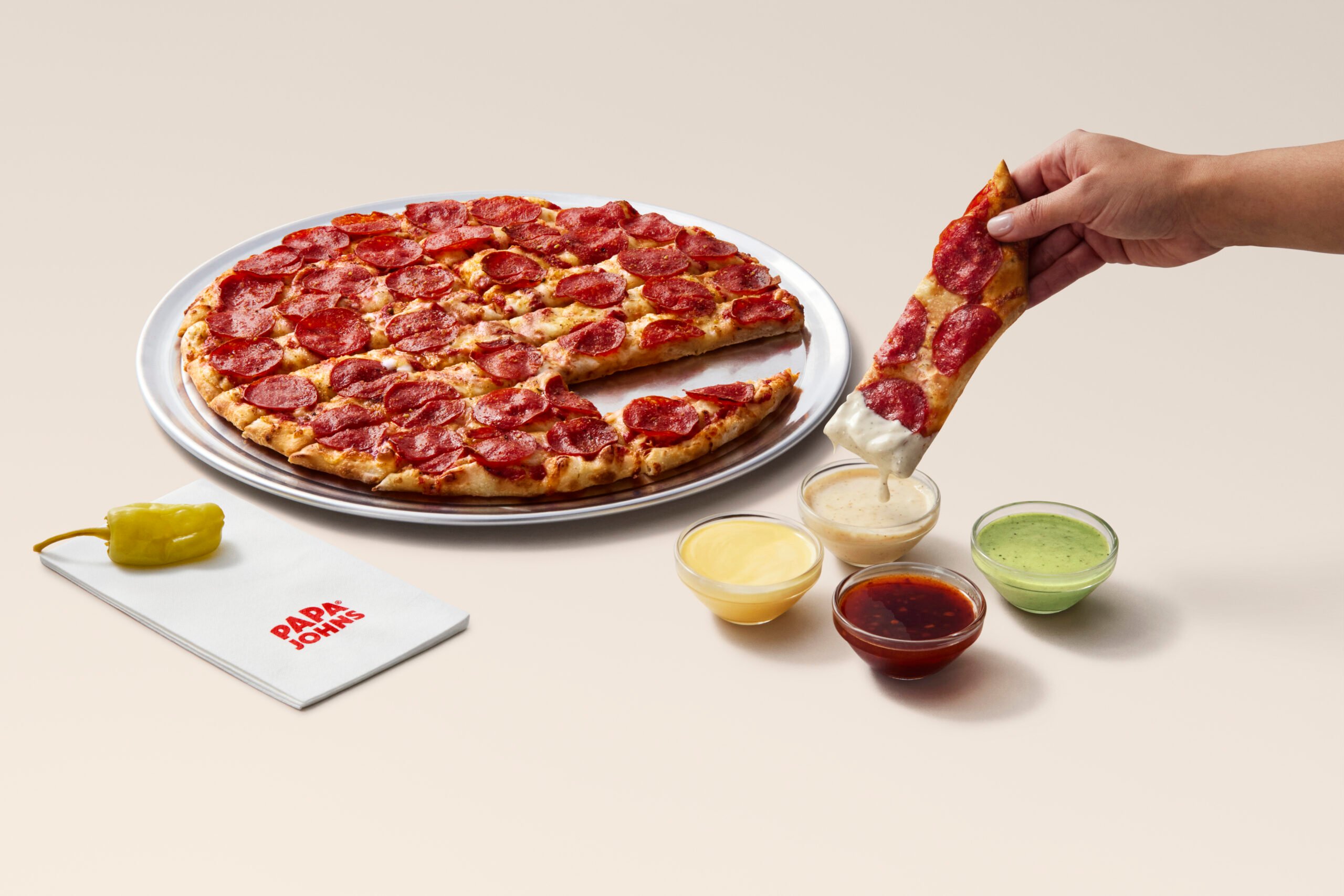 A hand dips a pepperoni pizza slice into a cup of creamy sauce. The new fast-food pizza sits on a silver tray with three dipping sauces, a pepperoncini, and a Papa Johns napkin nearby, all on a light background.