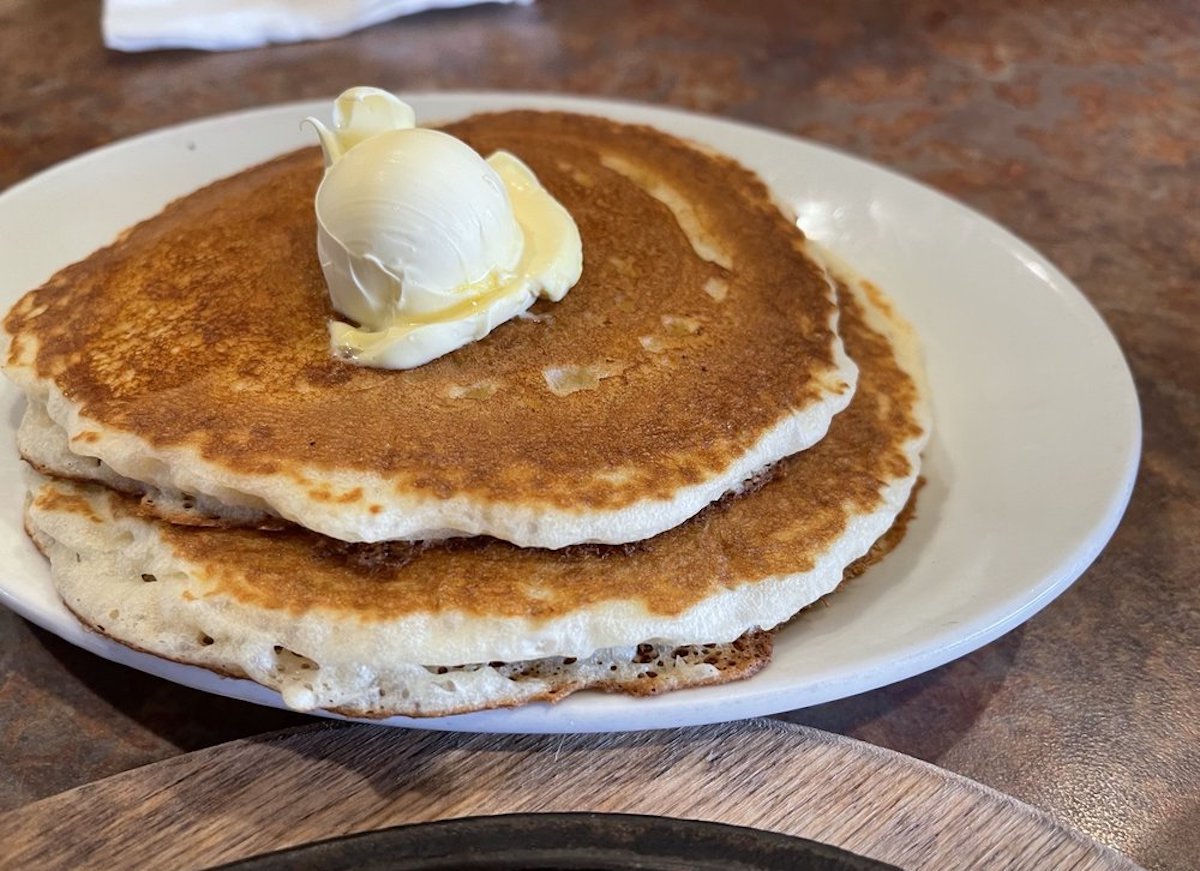 Two golden-brown pancakes stacked on a white plate, topped with a round scoop of butter starting to melt. The plate sits on a brown tabletop.