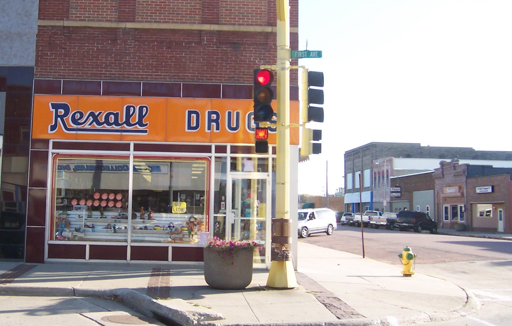 A corner street view featuring a Rexall Drug store with an orange sign, a traffic light, and a fire hydrant. Several buildings and parked cars line the street in the background.