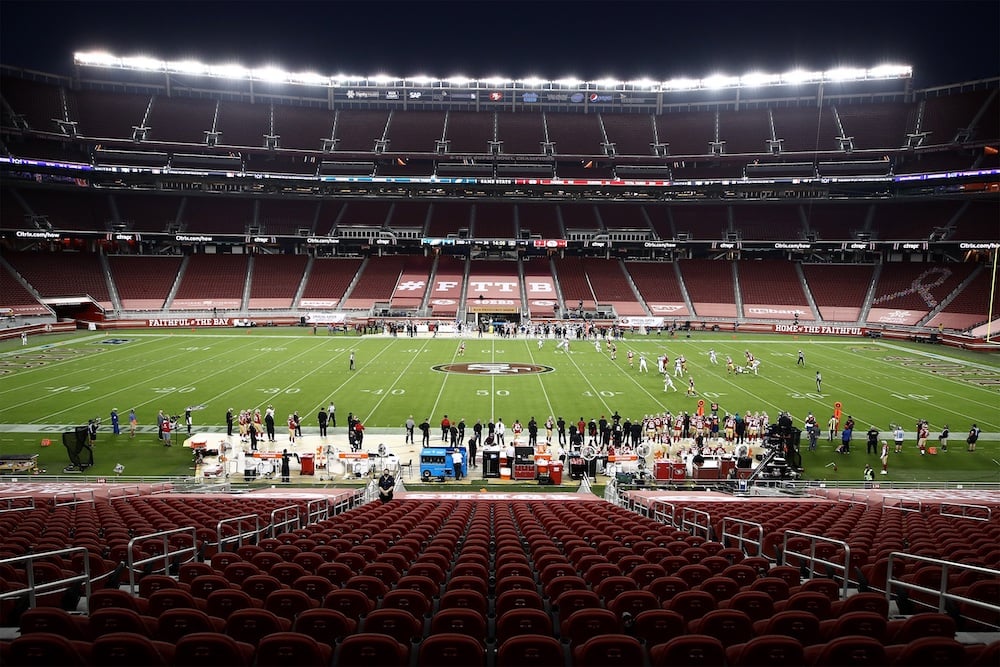 A nearly empty football stadium at night with players on the field and coaches on the sidelines. The seats are mostly vacant, and bright stadium lights illuminate the field.