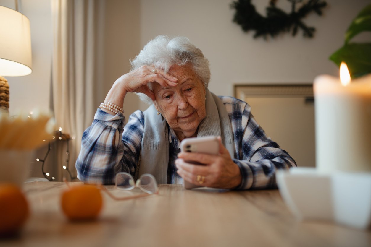 An elderly woman sits at a table, looking thoughtfully at her smartphone. Her glasses rest on the table nearby, and a candle, fruit, and a plant are in the foreground. Warm indoor lighting creates a cozy atmosphere.