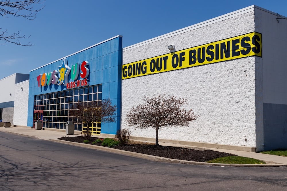 A Toys “R” Us store with a large “Going Out of Business” banner on the front. The building is mostly white with a blue facade, and the parking lot and surrounding trees are empty.