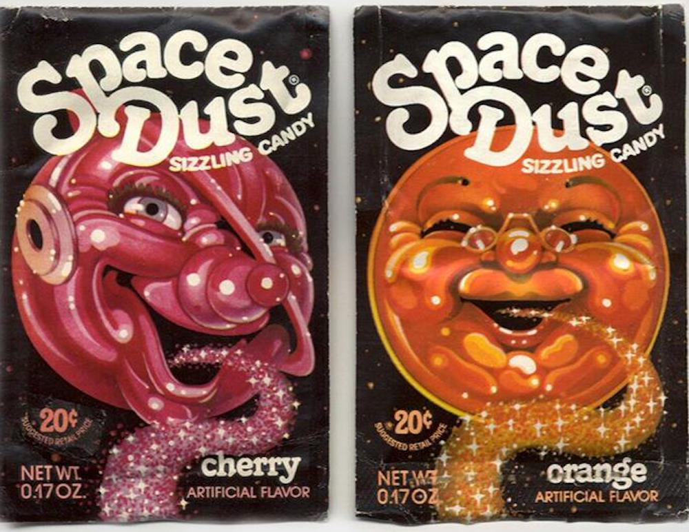Two vintage "Space Dust" candy packets: one cherry flavor with a smiling red cartoon face in a space helmet, and one orange flavor with a happy orange cartoon face, both with sparkling candy pouring from their mouths.