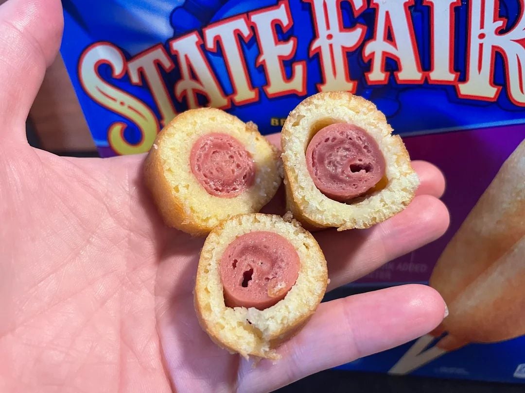 A hand holds three mini corn dogs with bites taken, showing the hot dog centers and cornbread coating. A blue "State Fair" food package is partially visible in the background.