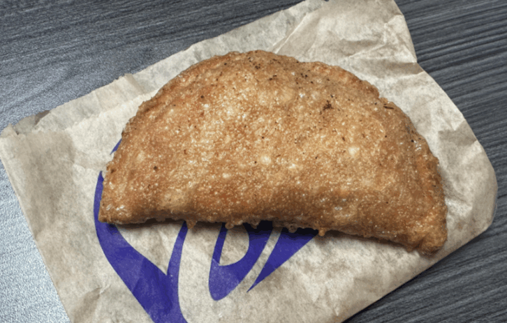Taco Bell's Caramel Apple Empanada, A golden-brown, crispy fried pastry sits on a piece of brown paper with a purple Taco Bell logo, placed on a dark wooden surface.
