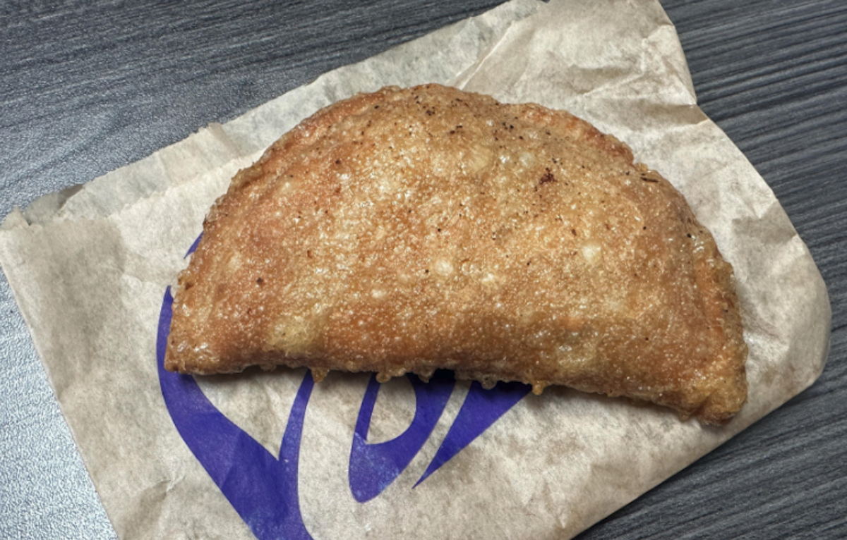 Taco Bell's Caramel Apple Empanada, A golden-brown, crispy fried pastry sits on a piece of brown paper with a purple Taco Bell logo, placed on a dark wooden surface.