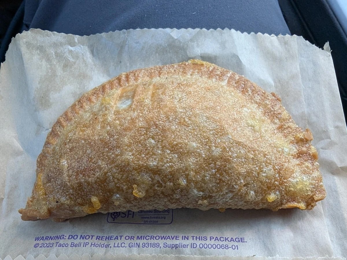 A golden-brown fried Taco Bell empanada sits on a paper wrapper, showing a crispy, flaky texture with some melted filling visible at the edges.