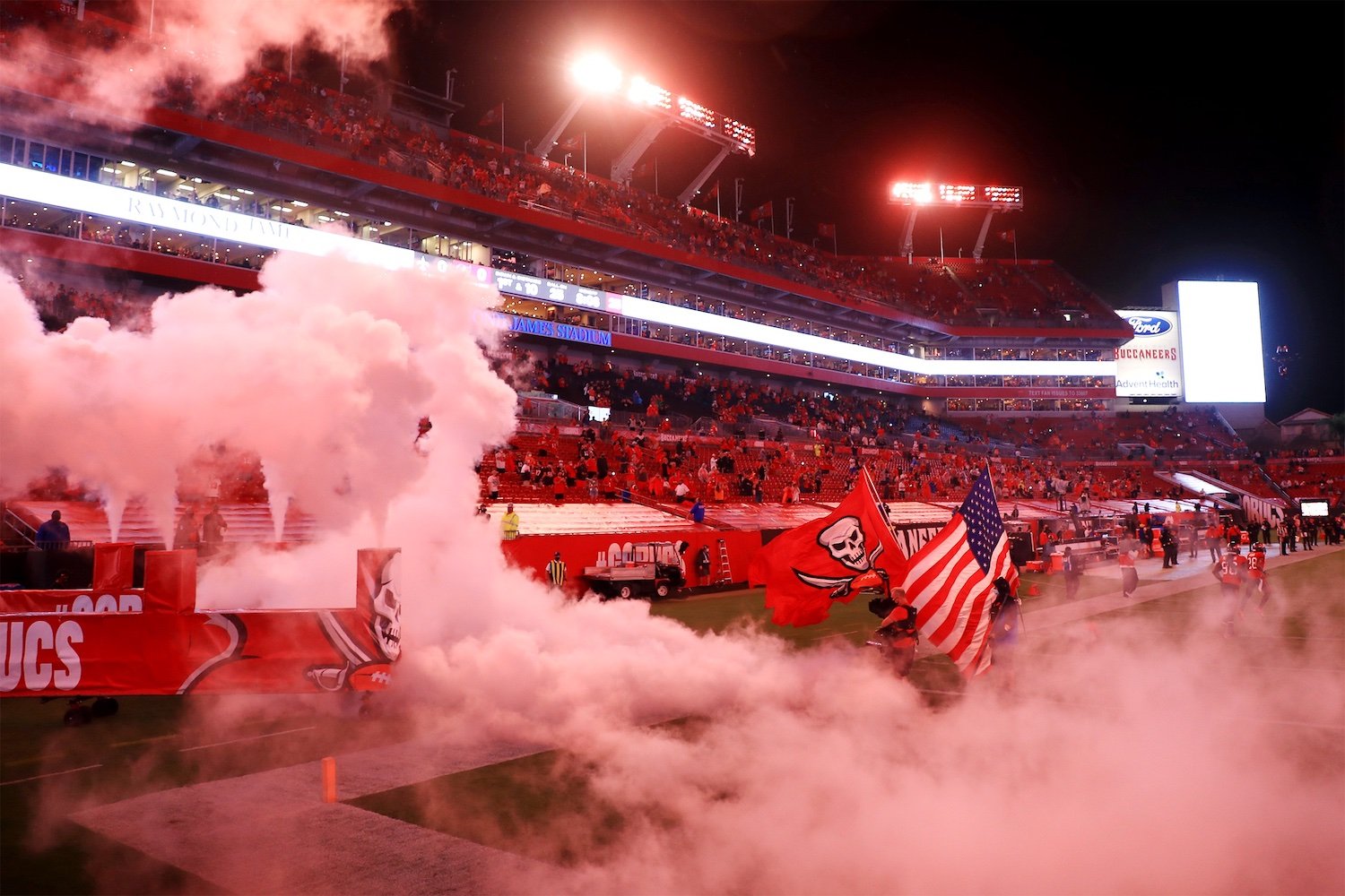 Players run onto a football field through smoke, carrying a Tampa Bay Buccaneers flag and an American flag, with stadium lights and fans in the red stands in the background at night.
