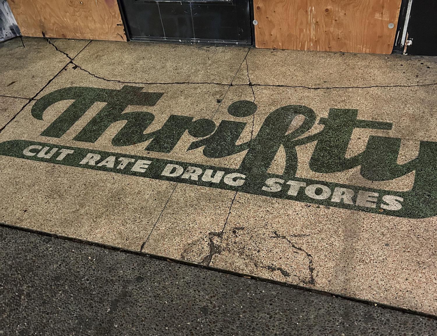 A faded green and white sidewalk sign reads "Thrifty Cut Rate Drug Stores" on cracked concrete in front of boarded-up storefronts.