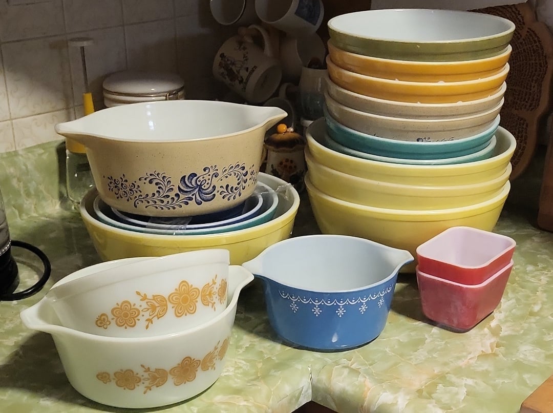 A collection of vintage mixing bowls and casserole dishes with decorative patterns in various colors, stacked and arranged on a green marbled countertop in a kitchen.