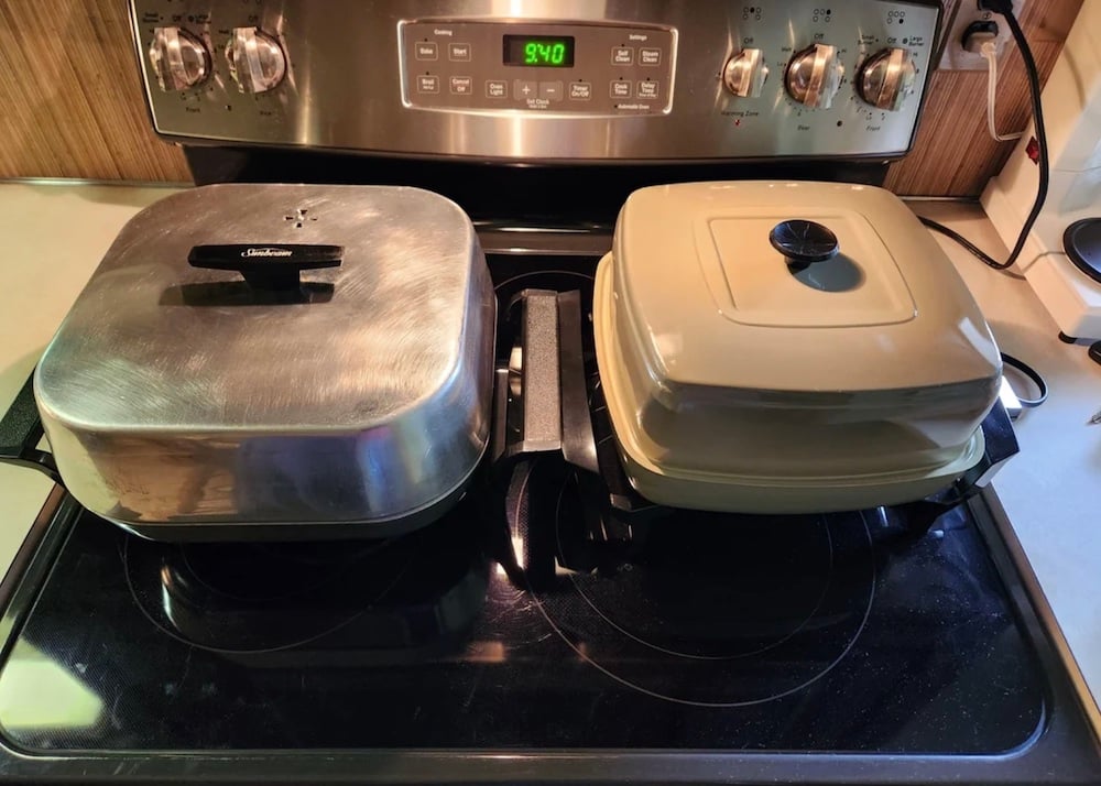 Two vintage closed electric skillets sit side by side on a black stove, one with a silver metal lid and one with a cream-colored lid. The stove display shows 9:40 and the knobs are set in various positions.