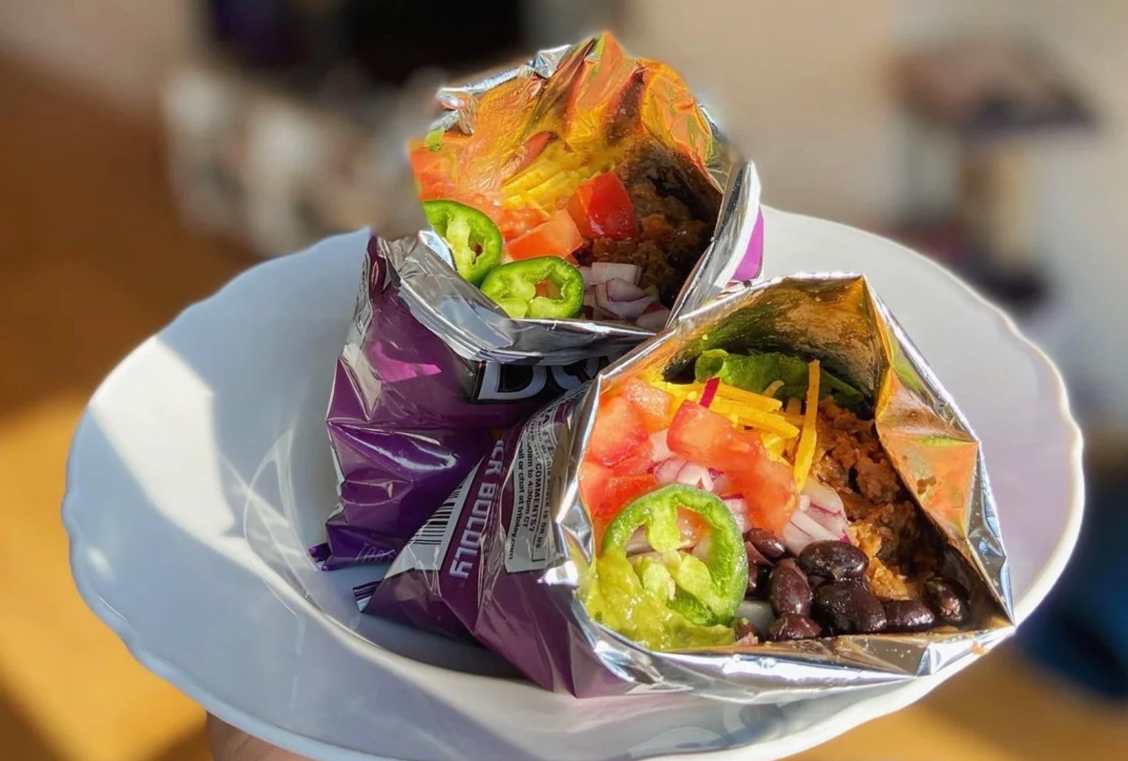 Two opened bags of Doritos on a white plate, each filled with taco ingredients like ground beef, black beans, shredded cheese, lettuce, diced tomatoes, onions, guacamole, and sliced jalapeños.