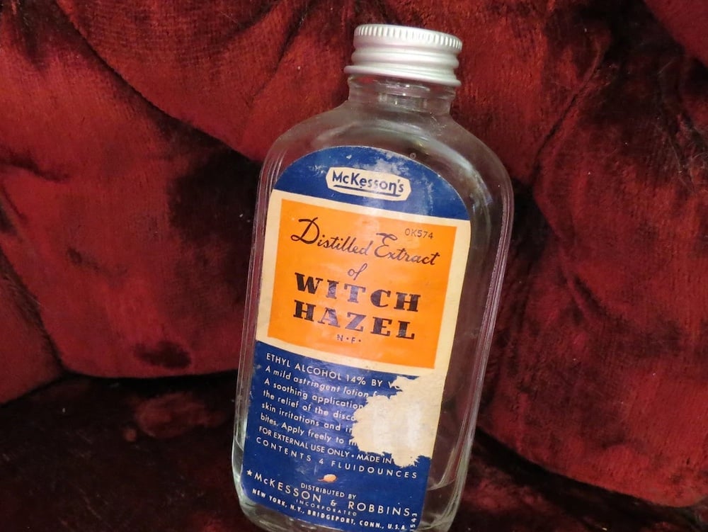 A clear glass bottle labeled "Distilled Extract of Witch Hazel" sits on a deep red, textured fabric. The blue and gold label notes the product is distributed by McKesson & Robbins of New York.
